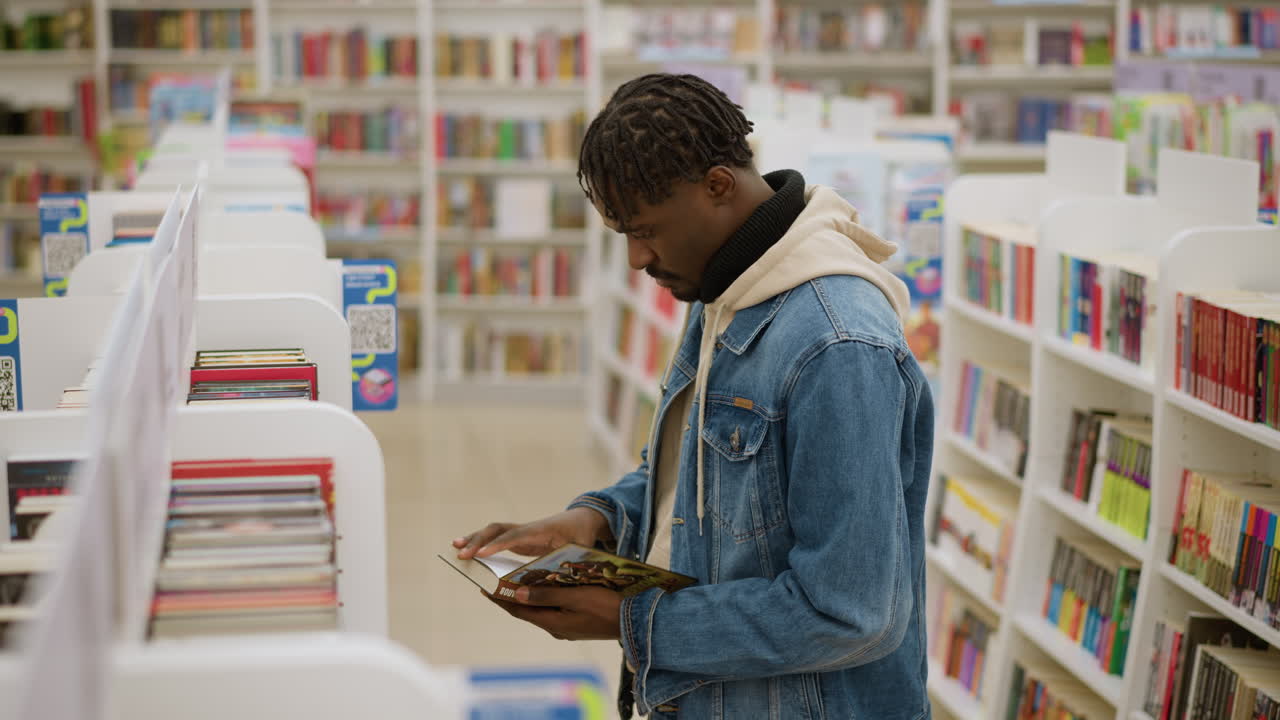 Big boy choosing books in modern library. Focused young man in denim jacket browsing bookshelves in bookstore surrounded by neatly organized colorful book spines, deep in thought while selecting