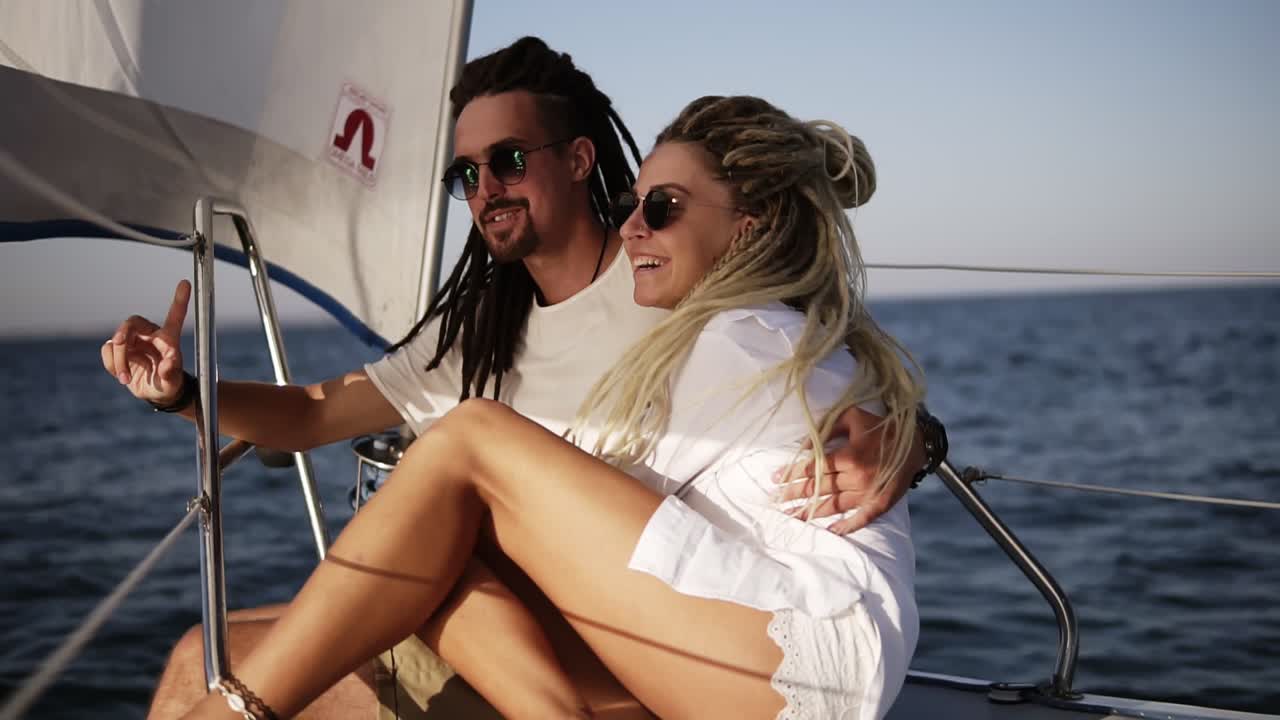 Stylish couple with dreadlocks in white clothes and sunglasses sitting embracing on the bow of the yacht and smiling. Loving