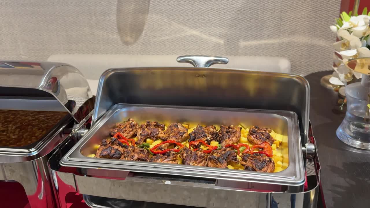 Allestimento buffet catering interno con scaldavivande in acciaio inossidabile contenente pezzi di pollo grigliato su patate e peperoni a fette, esposizione di cibo per linea di servizio sotto un'illuminazione calda da ristorante