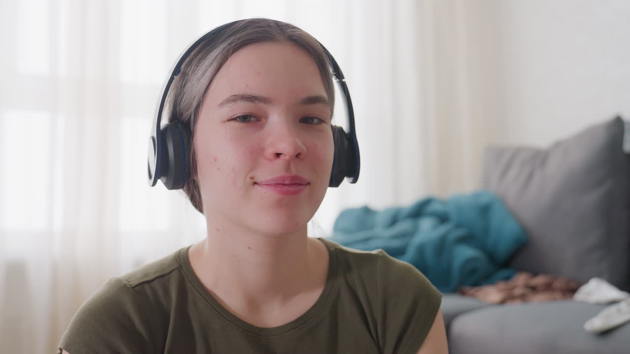 Close up of young woman with headphones nodding head to rhythm of music, calm expression showing enjoyment while seated indoors, natural daylight from window