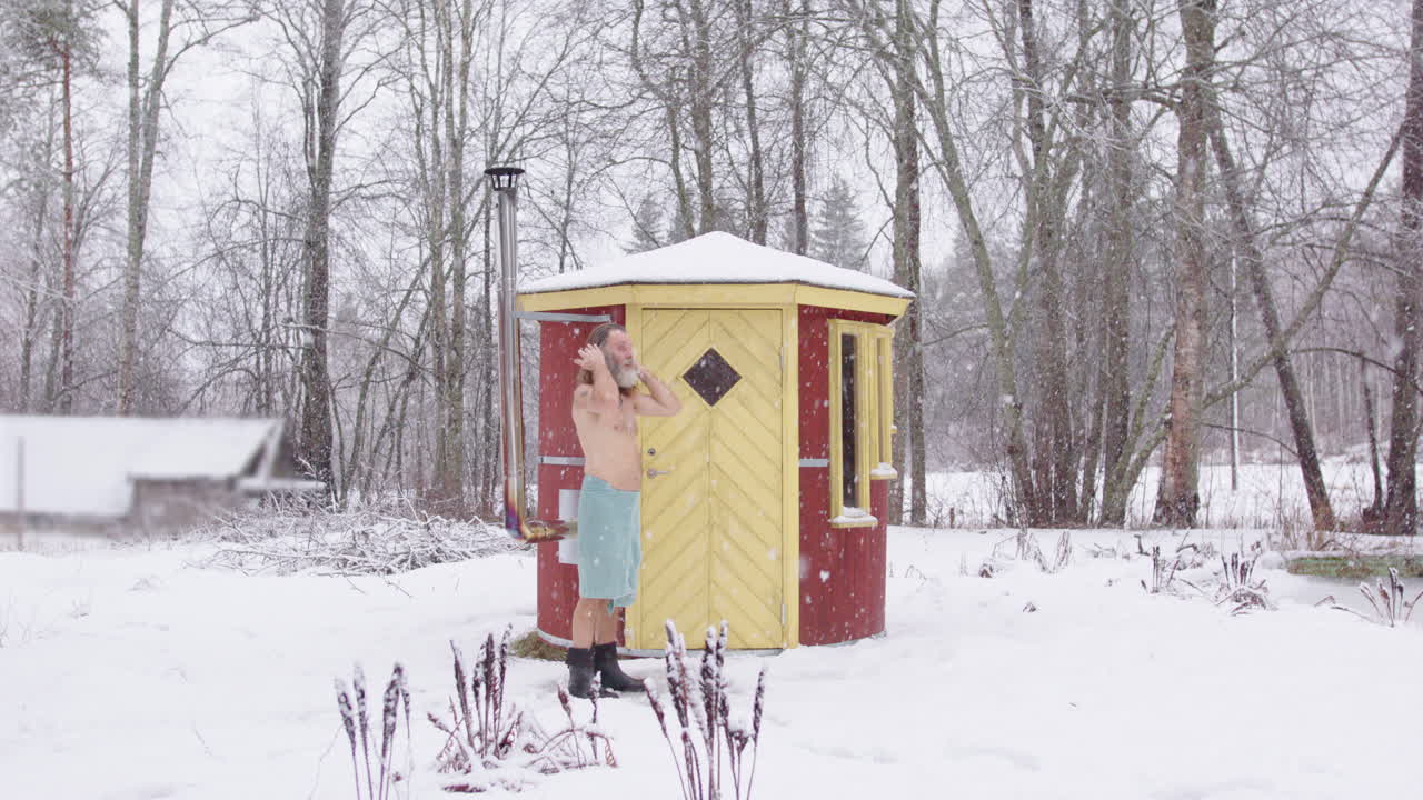 Hippy man bare chested in towel exits wooden sauna into snowy environment