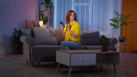 stay at home, joyful young housewife wearing glasses uses mobile phone while sitting on couch while relaxing in living room at night