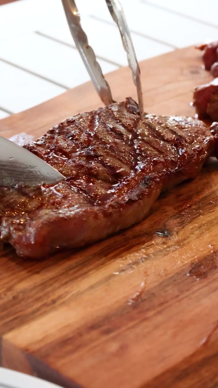 A steak is sliced on a wooden board using a knife and tongs, under natural lighting