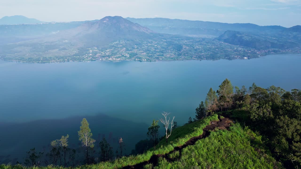 Early morning sunlight softly illuminates the green slopes of Trunyan Hill in Bali, captured in smooth drone footage with Mount Batur and the calm Lake Batur completing the stunning scene.