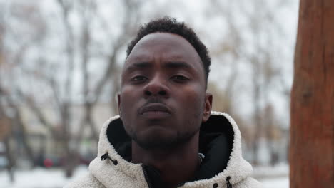 confident young man with bold expression wearing winter jacket, standing outdoors with blurred background featuring trees and unclear urban structures, exuding determination in cold weather