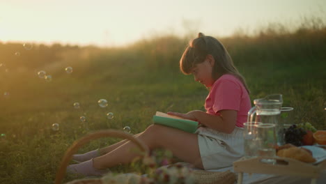 little girl sitting outdoors during golden hour reading book and turning page, surrounded by picnic setup with fruits, croissant, and bubbles gently floating in air, sunlight illuminating background