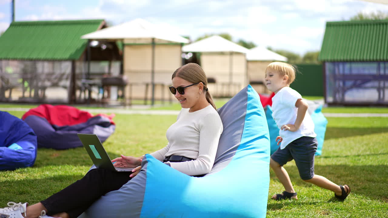 Mother and her son playing outdoors. Young woman distracts from work on laptop to pay attention to her child. Summer time leisure in rest zone. Blurred backdrop.
