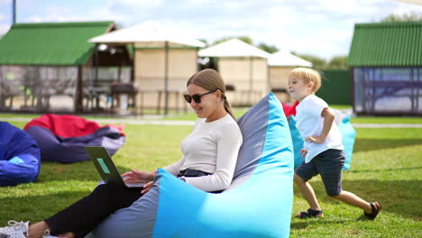 mother and her son playing outdoors. young woman distracts from work on laptop to pay attention to her child. summer time leisure in rest zone. blurred backdrop.