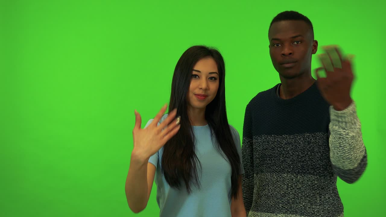 A young Asian woman and a young black man wave at the camera in a gesture of invitation - green screen studio