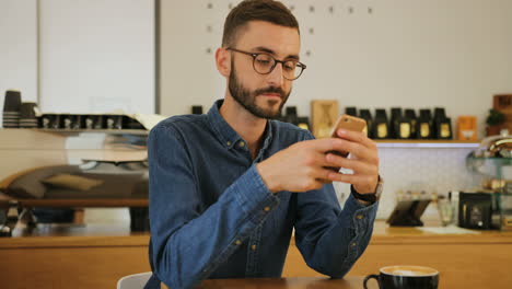 young caucasian man in glasses using smartphone while sitting at the table in a coffee shop