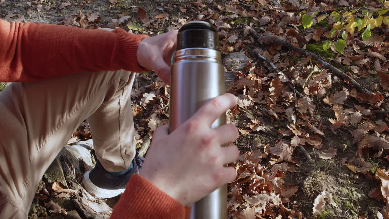 hombre sirviendo café o té caliente de una botella de termo en una taza en el bosque, hojas de otoño en el fondo