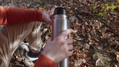 Man-pouring-hot-coffee-or-tea-from-thermos-bottle-into-mug-in-forest,-autumn-leaves-in-background