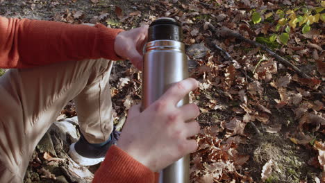 man pouring hot coffee or tea from thermos bottle into mug in forest, autumn leaves in background