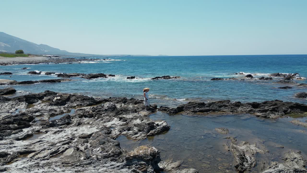 Lone Female Tourist Standing On Rocky Coastline In Island Of Crete In Greece. orbiting drone shot