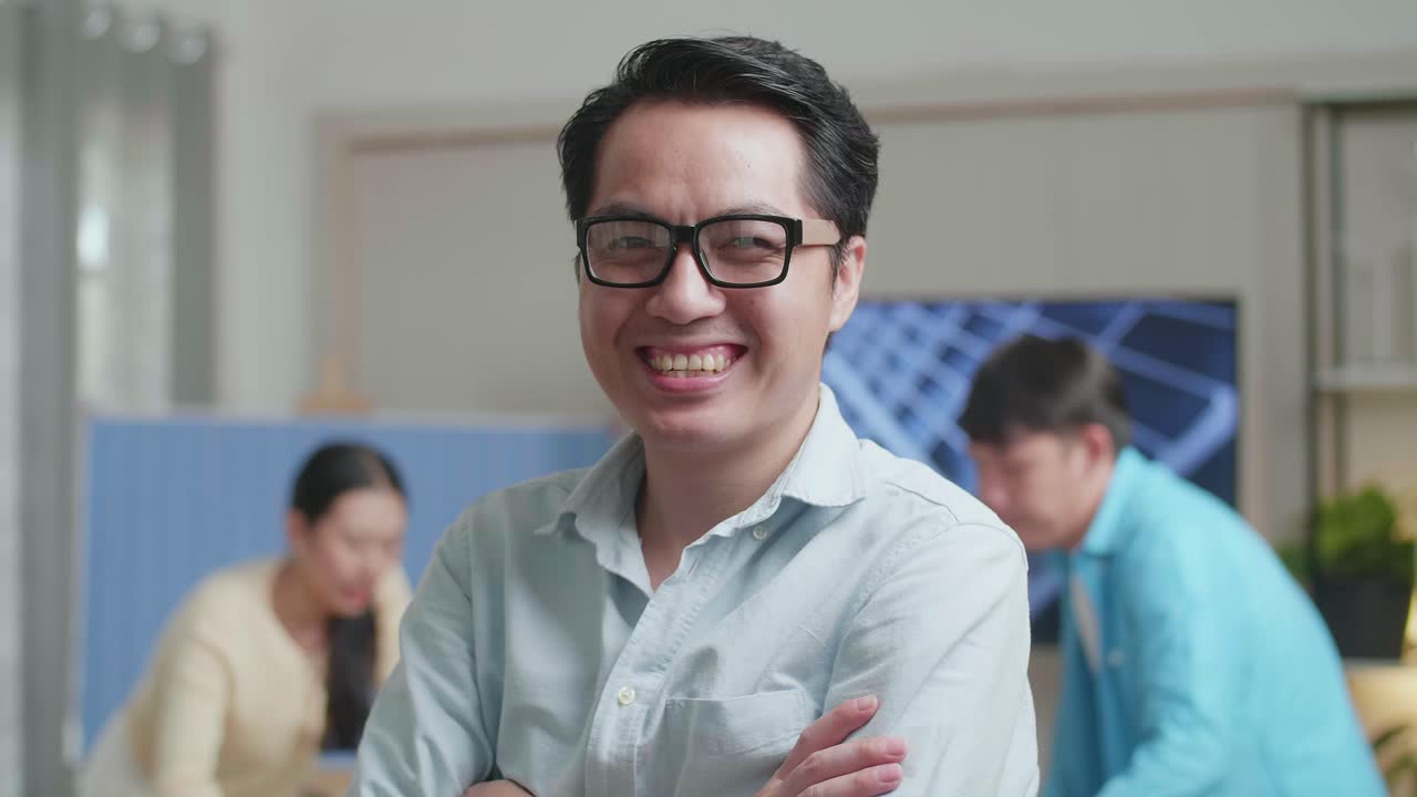 미소 짓는 아시아 남성 엔지니어가 동료들이 사무실에서 태양전지 작업에 대해 논의하는 동안 카메라에 팔을 고 있습니다.