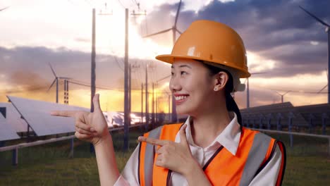 close up of asian female engineer with safety helmet smiling and pointing to side with solar panel and wind turbines