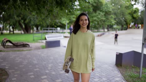 A-happy-brunette-girl-in-a-green-sweater-walks-with-a-skateboard-in-the-park-and-smiles.-Walk-in-the-park,-portrait.-Hobby