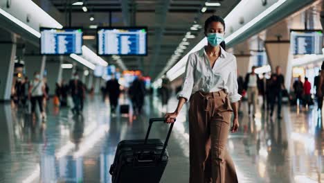 elegant businesswoman wearing protective face mask is walking through airport terminal pulling suitcase, ready for business trip during pandemic