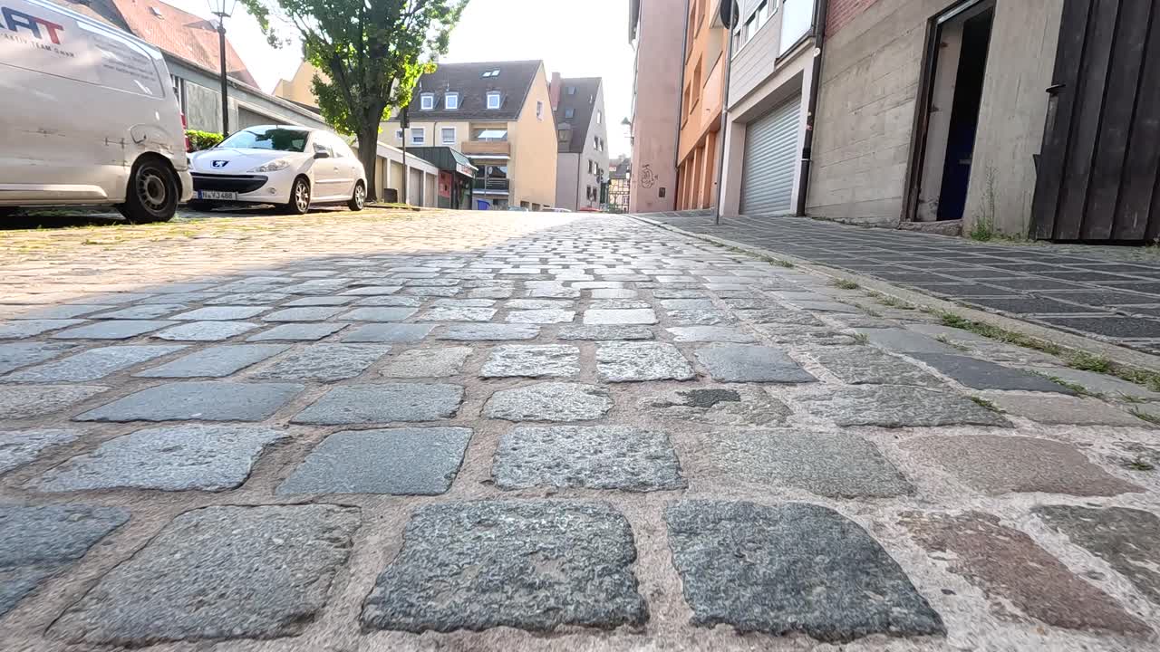 Low-angle tracking shot along sunlit cobblestone alley with parked cars and historic buildings, Nuremberg