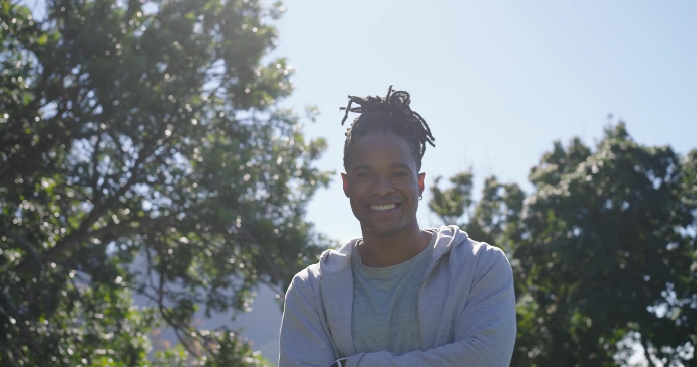 African American man hoodie-clad walking by hedge in park looking at trees, touching hedge smiling