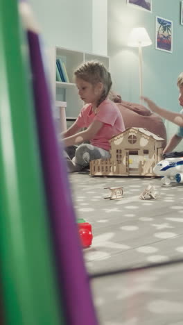 children make structures from plastic constructor blocks sitting near dollhouse. siblings play with toys view through clearance in bunch of pencils in glass