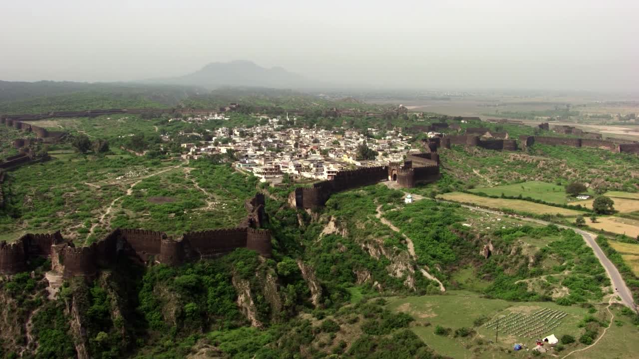 An aerial view shows an ancient fortified town on a hilltop in Pakistan. The expansive stone fortifications with large bastions encircle a dense cluster of white and beige buildings