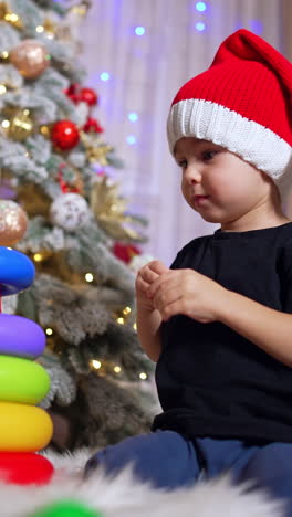 toddler boy in black t-shirt playing with toy pyramid. kid wearing santa cap assembling toy near the christmas tree. low angle view. vertical video