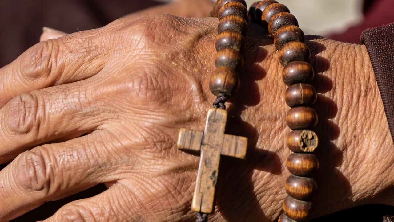 Mãos de idoso se aproximando, dedo deslizando terço de madeira e cruz para oração, manga bordô.