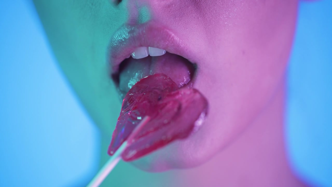 Slow motion 4K, donna che lecca un lecca-lecca rosa a forma di cuore, illuminazione al neon, tenue bagliore rosa e verde sul viso, sfondo blu, colori vivaci, atmosfera giocosa, bellezza cinematografica, atmosfera dolce.