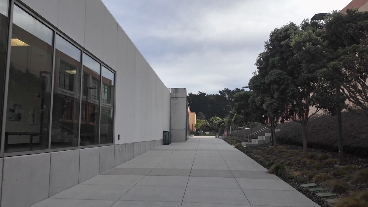 Pov shot walking along a concrete pathway between two modern buildings at Skyline College in San Bruno, California, on a cloudy day, showing the campus architecture and landscaping