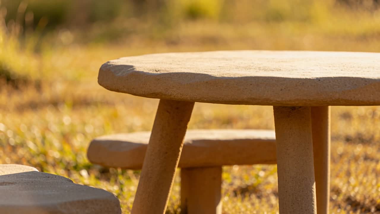 Starting closeup, camera pulling back revealing stone table with angled legs and stools on grass