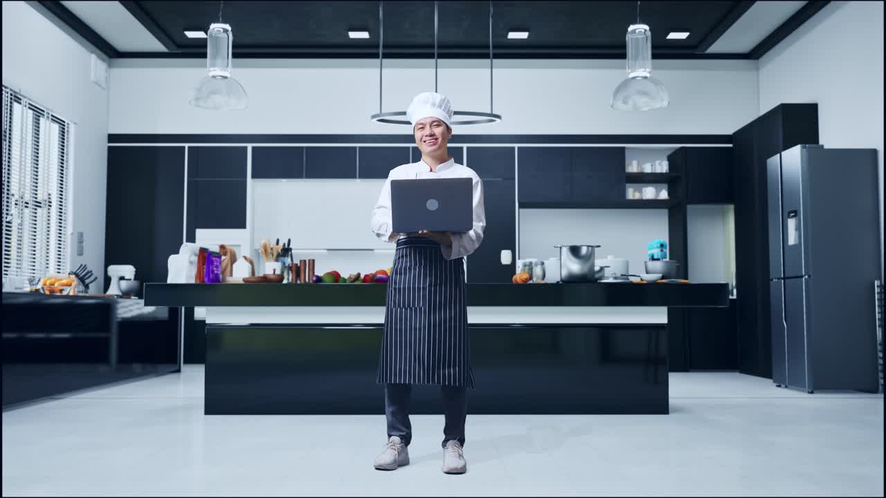 cuerpo lleno de cocinero hombre asiático usando una computadora portátil y sonriendo a la cámara mientras está de pie en la cocina de casa