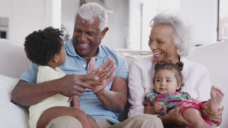 grandparents sitting on sofa at home playing game with baby granddaughters