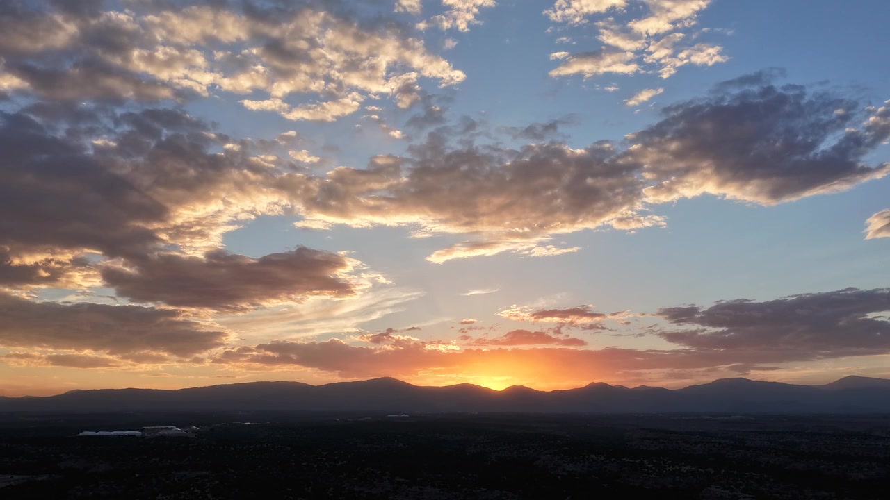 4K UHD drone timelapse capturing a dreamy sunset. Soft pastel colors shift dynamically from behind a dark mountain range silhouette, creating a serene, colorful atmospheric sky.