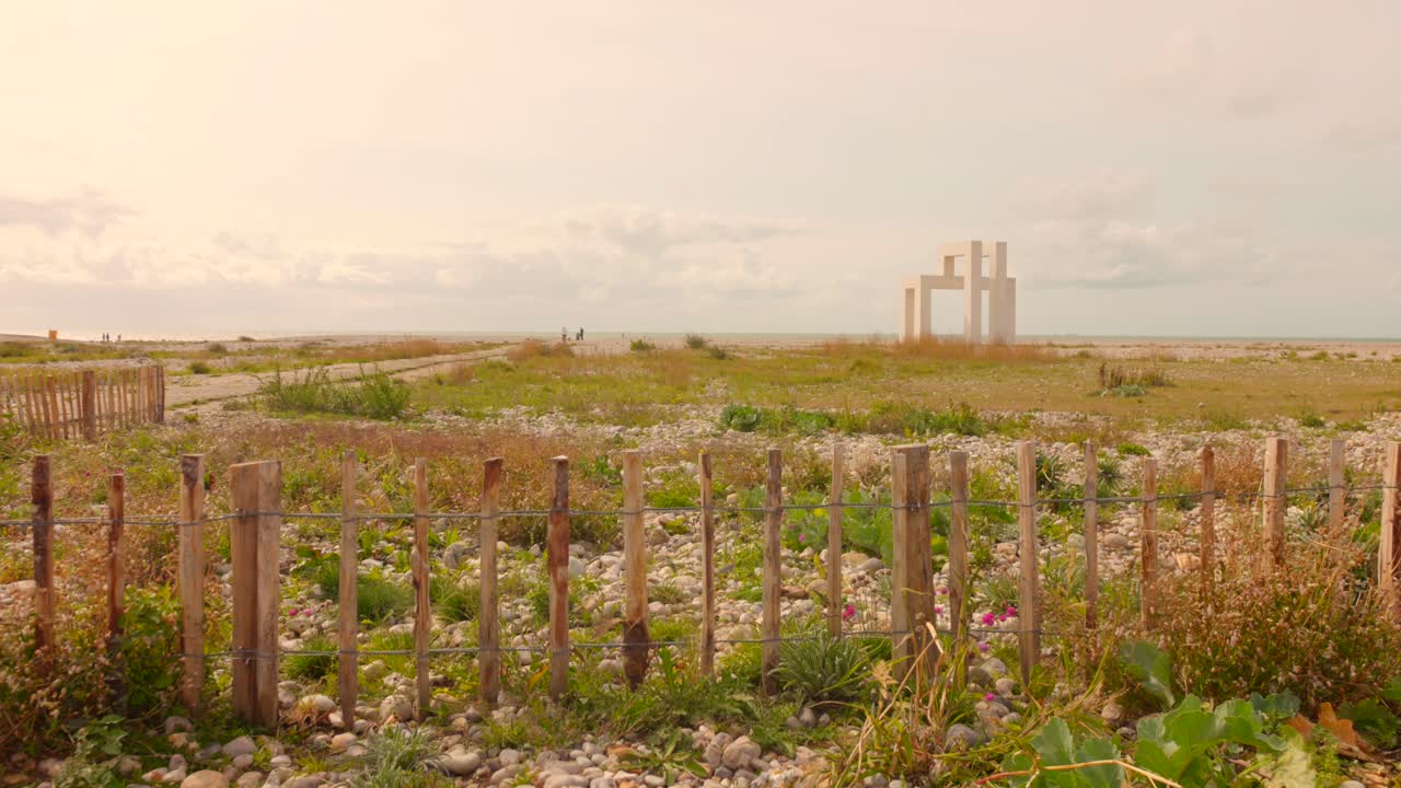 Ampia inquadratura cinematografica della spiaggia di Le Havre all'ora d'oro, con pali di recinzione in legno stagionato, dune di fiori selvatici costieri e iconici edifici modernisti all'orizzonte.