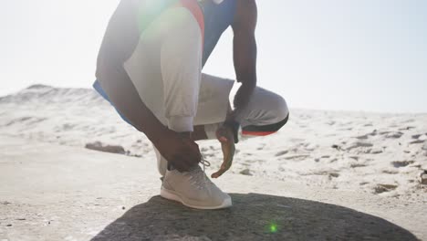 Midsection-of-african-american-man-tying-shoelaces-during-exercise-outdoors-on-beach