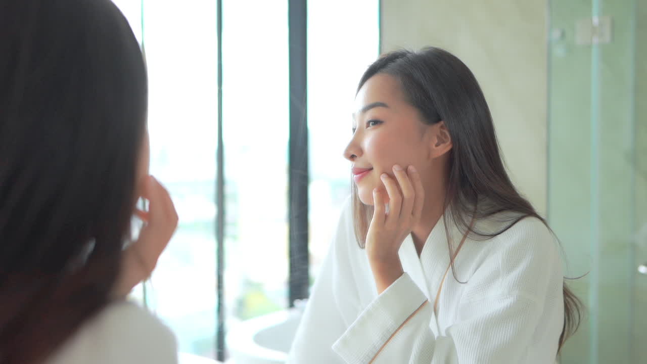 목욕 가운을 입은 아시아 여성은 욕실 슬로모션 아침에 웃는 거울 근처에서 얼굴 피부를 확인하고 만집니다.