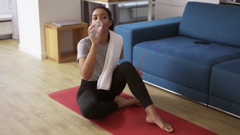 mujer afroamericana deportiva bebiendo agua después del entrenamiento en casa en un tapete de yoga