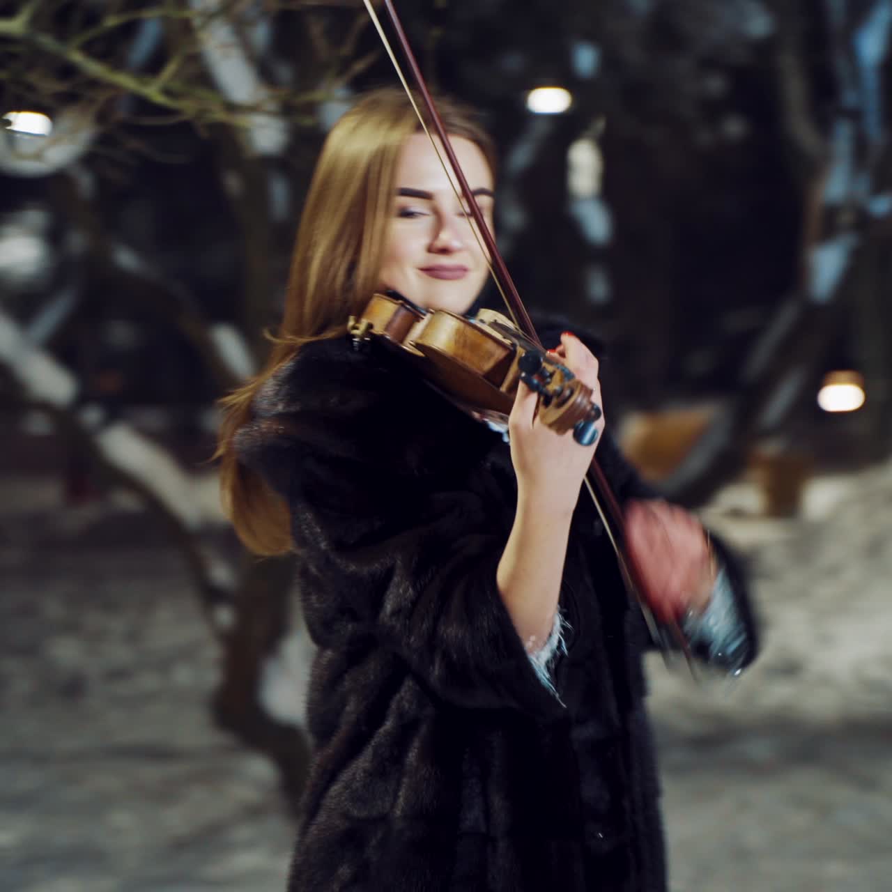 A talented girl in warm clothes is performing composition on the violin in a winter park in the evening on a blurred background. Camera motion to right