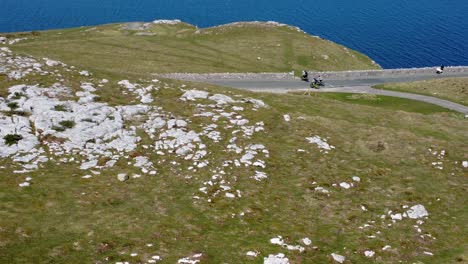 tourists riding mountain bike along scenic green mountain country road overlooking gorgeous blue irish sea aerial rising dolly left