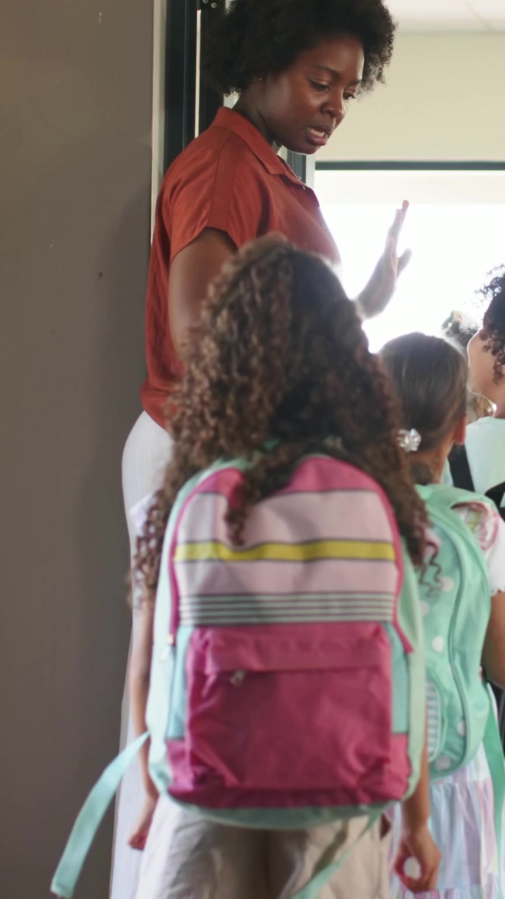 Vertical video: Walking kid with bag entering school as teacher in rust top waving, stepping aside
