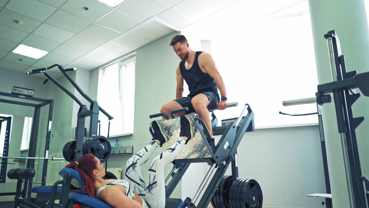 Sporty girl doing exercise for legs on a modern simulator in the gym. Woman is lifting man by her strong legs on a fitness machine in the light sports center.