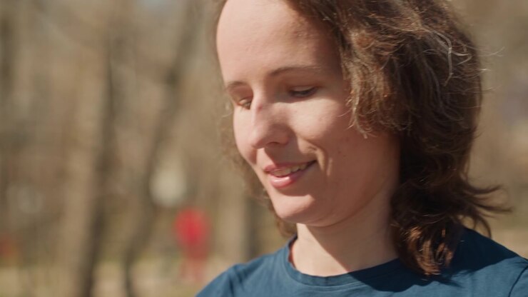 Primo piano di una donna che parla all'aperto con luce naturale, le sue espressioni visibili mentre comunica. Lo sfondo mostra alberi sfocati e la luce del giorno in un ambiente esterno, suggerendo un clima sereno.