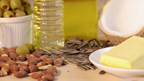 a sunflower seed falls onto a plate of butter surrounded by almonds, olives, sunflower seeds, coconut, and a bottle of oil in bright studio lighting