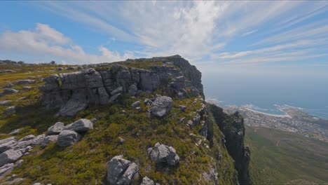 Toma-Aérea-Volando-Sobre-La-Naturaleza-Escarpada-En-La-Montaña-De-La-Mesa-En-Sudáfrica,-Con-Ciudad-Del-Cabo-Y-El-Océano-Muy-Por-Debajo-En-La-Distancia