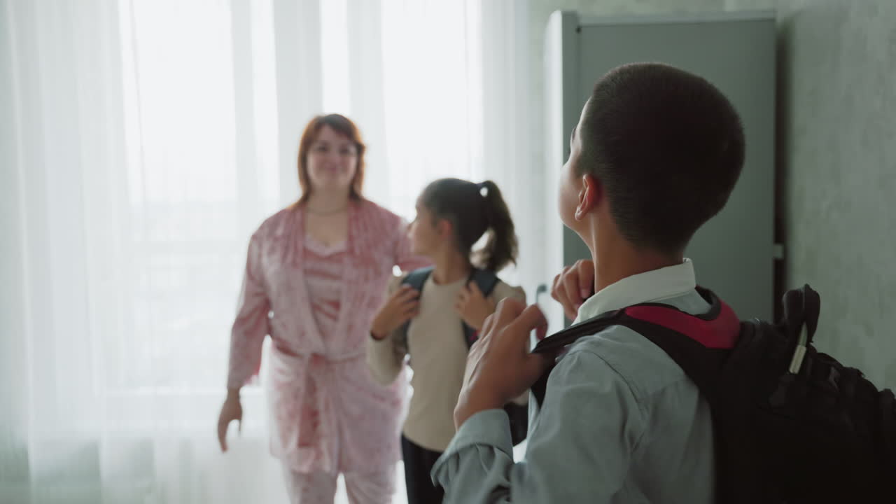 kids in casual clothes gathering morning supplies for school in sunlit kitchen, boy adjusts backpack straps while girl packs textbooks with mother nearby offering guidance, warm familial scene