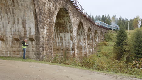 man inspecting a stone arch bridge with a train passing by