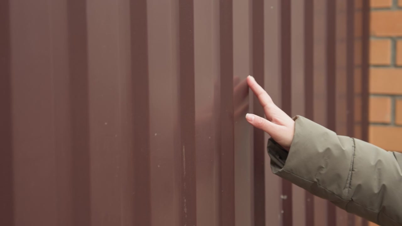 Caucasian woman touches brown metal fence near brick pillar, winter coat and glasses, homebuyer inspecting property boundary with cautious fingertips, closeup texture study, muted overcast street