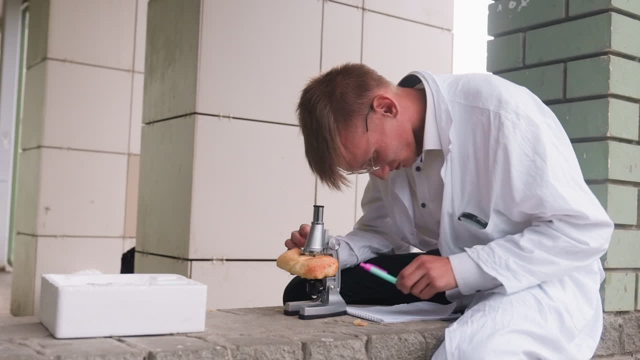 Side view of scientific researcher in white coat carefully examining pastry through microscope in outdoor setting, reflecting focus, curiosity, and scientific observation while jotting with pen