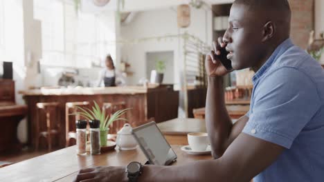 Hombre-Afroamericano-Hablando-Por-Teléfono-Inteligente-Y-Usando-Tableta-En-La-Cafetería,-Cámara-Lenta